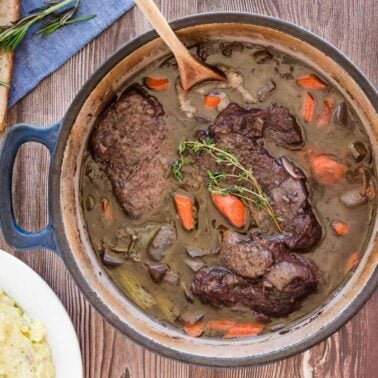 A pot of Southern pot roast with carrots and herbs, placed on a wooden table next to a loaf of bread and a bowl of mashed potatoes.