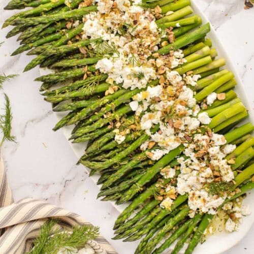 A platter of green asparagus topped with crumbled feta cheese, chopped nuts, and herbs, on a marble surface, next to a striped cloth napkin.