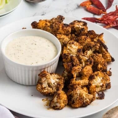 Roasted cauliflower bites with a side of creamy dip on a white plate, accompanied by dried red chilies and cucumber slices in the background.