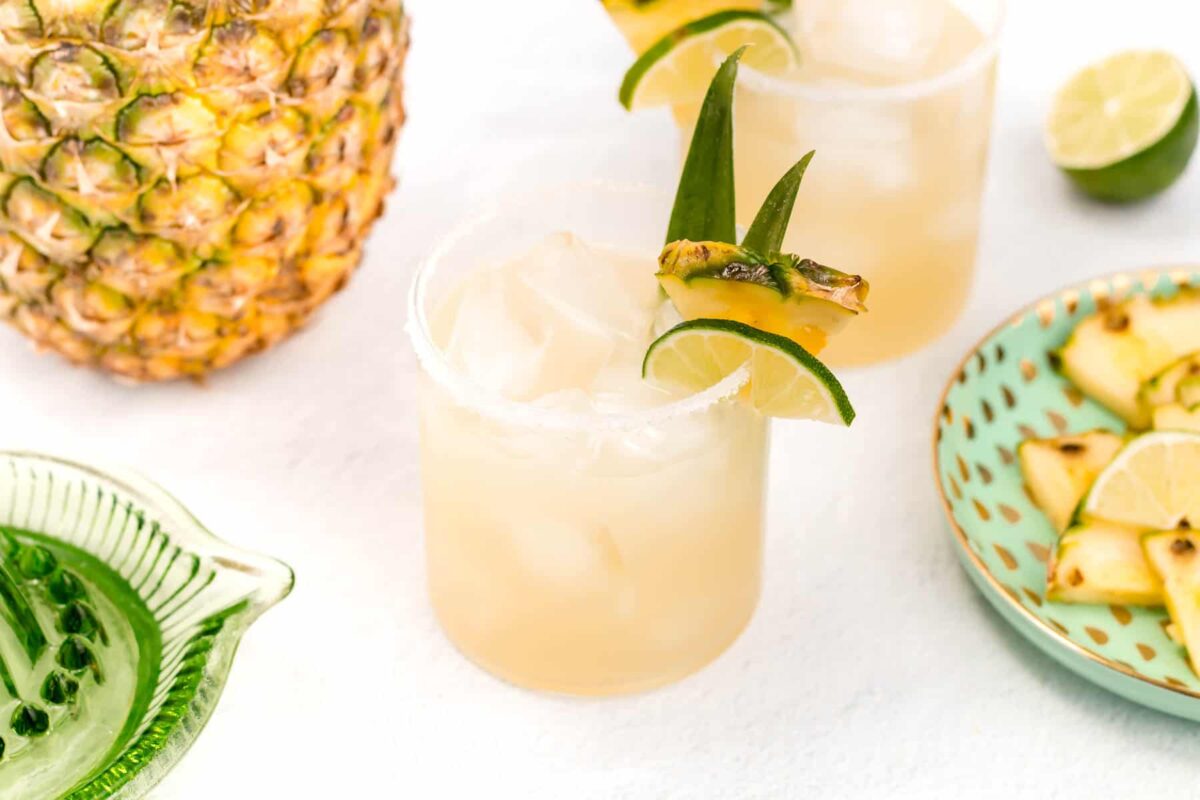 A glass of pineapple cocktail with ice, garnished with a pineapple slice and lime, sits next to a fresh pineapple, lime halves, and a plate of pineapple slices—a must-try party drink inspired by top Margarita recipes.