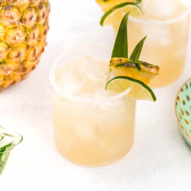 A glass of pineapple cocktail with ice, garnished with a pineapple slice and lime, sits next to a fresh pineapple, lime halves, and a plate of pineapple slices—a must-try party drink inspired by top Margarita recipes.