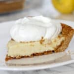 A slice of lemon icebox pie with a creamy filling and graham cracker crust, topped with a large dollop of whipped cream on a white plate.