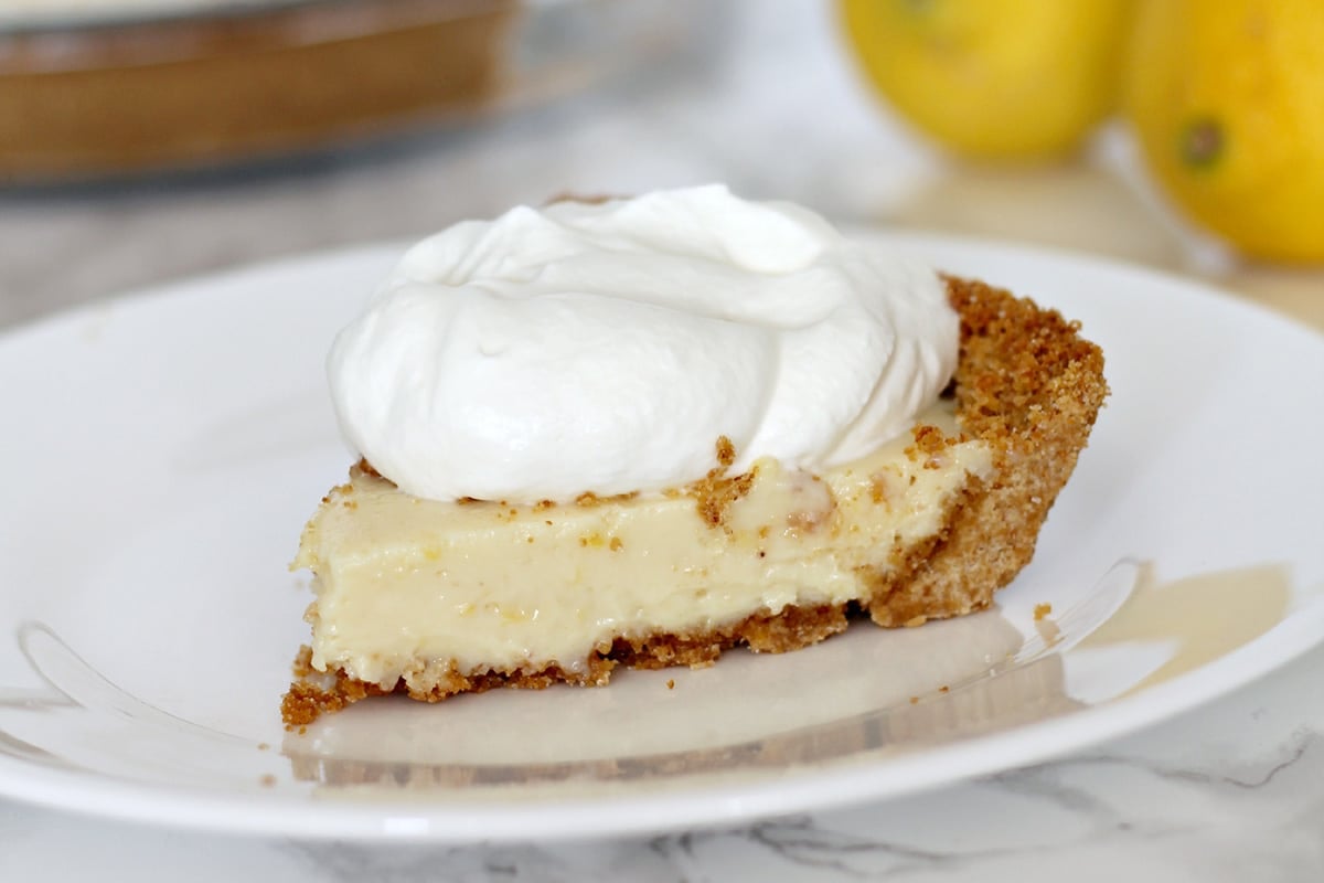 A slice of lemon icebox pie with a creamy filling and graham cracker crust, topped with a large dollop of whipped cream on a white plate.