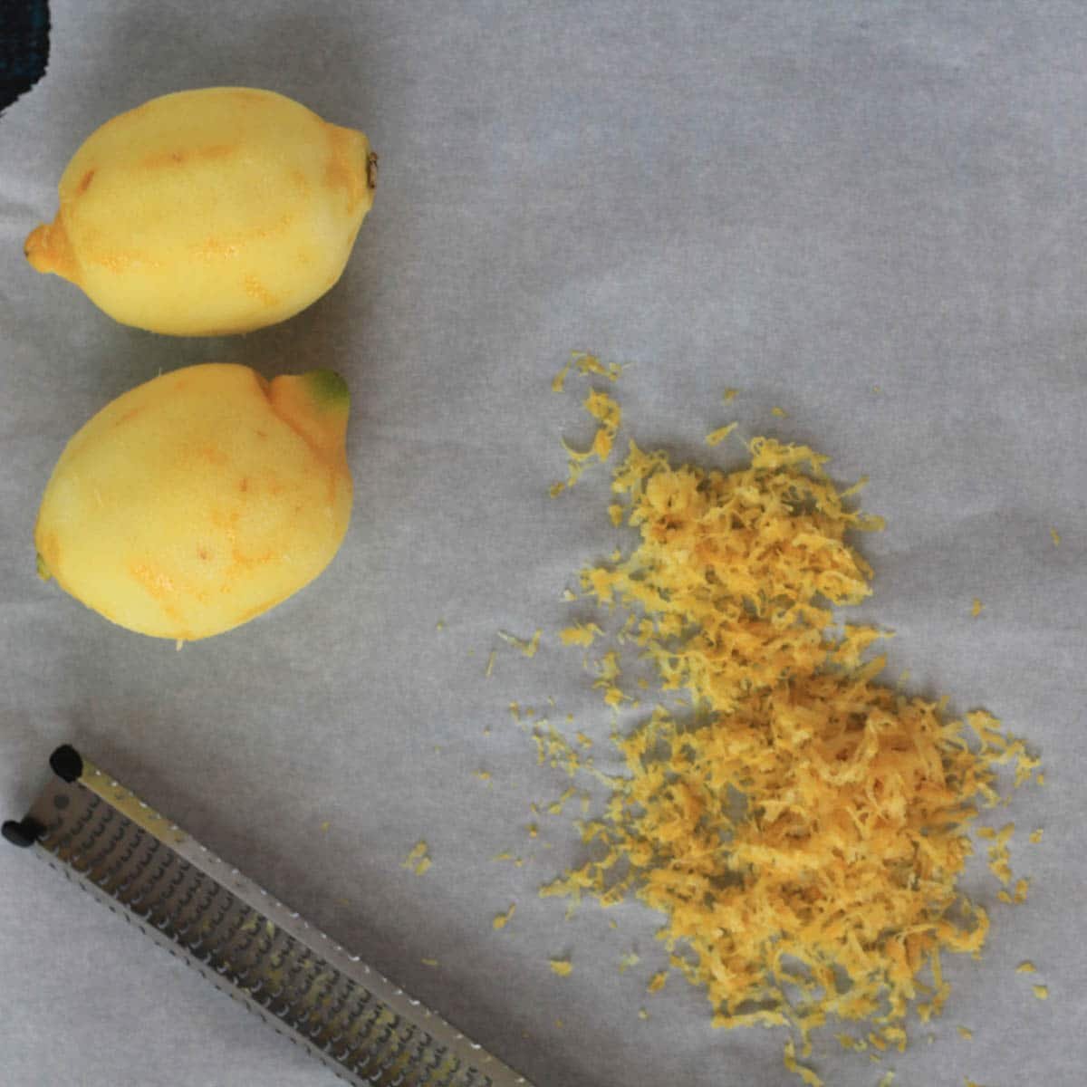 Two whole lemons, a pile of lemon zest, and a metal grater are placed on a sheet of parchment paper.