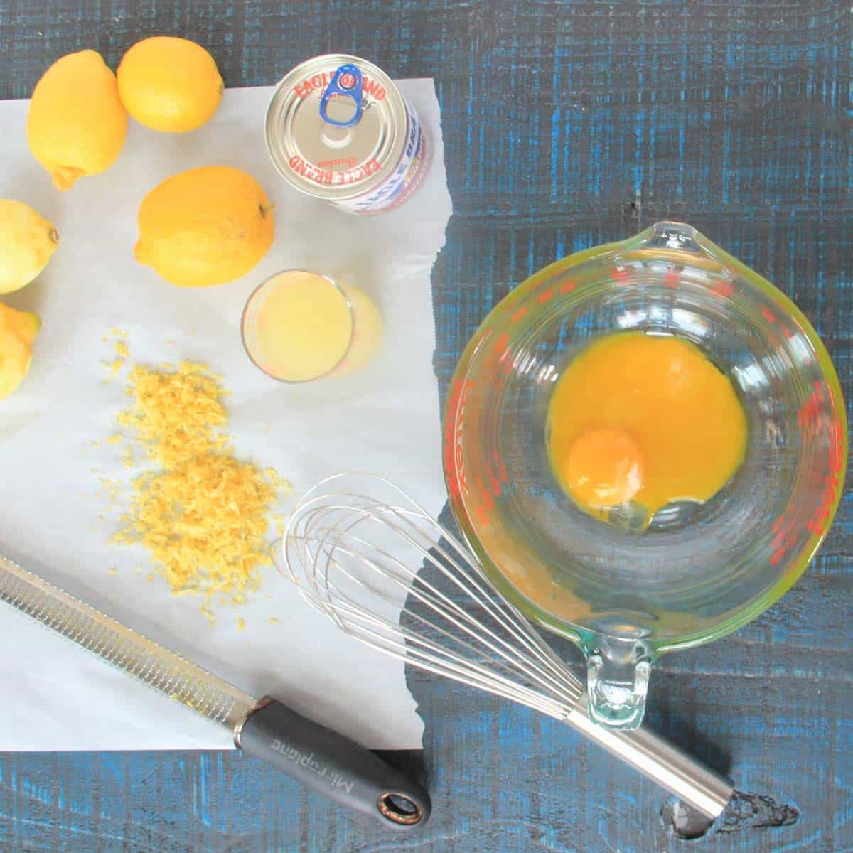 Overhead view of lemons, lemon zest, a can of sweetened condensed milk, lemon juice, a whisk, and eggs in a glass measuring cup on a dark surface.
