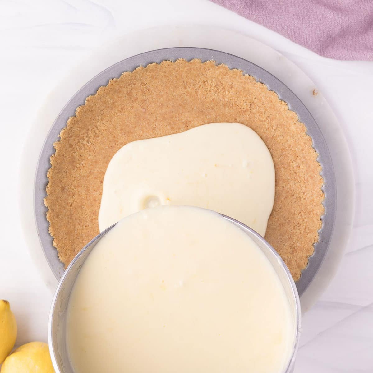A metal bowl pours lemon pie filling onto a graham cracker crust in a round pan, with lemons and a purple cloth nearby.