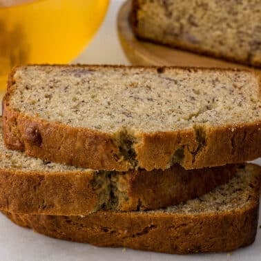 Three slices of sour cream banana bread stacked on a table with a loaf and a honey container in the background.