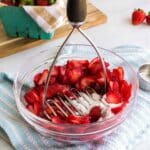A bowl of sliced strawberries being mashed with sugar using a potato masher. A bunch of strawberries sits in a basket in the background.