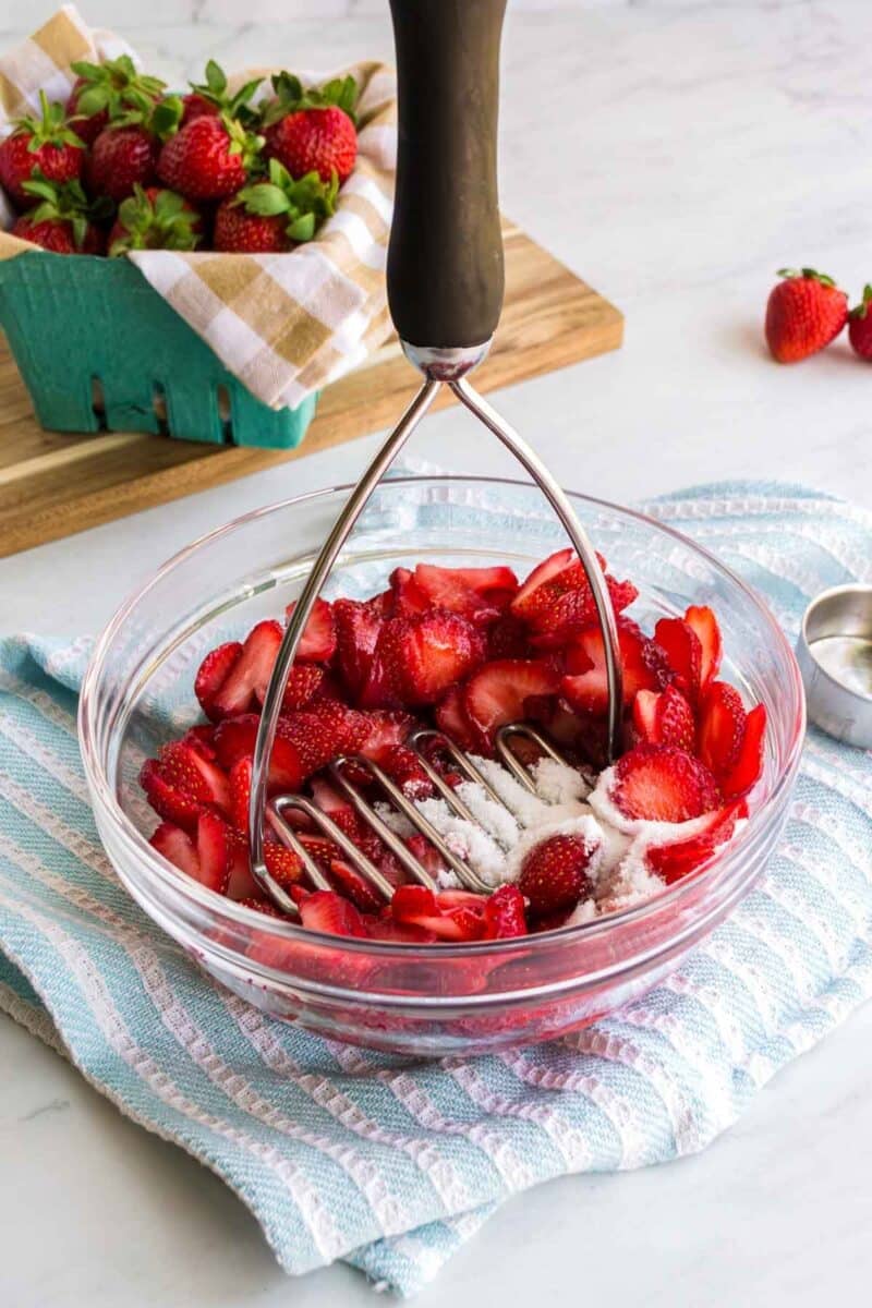 A bowl of sliced strawberries being mashed with sugar using a potato masher. A bunch of strawberries sits in a basket in the background.