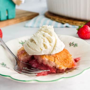 A plate with strawberry cobbler topped with a scoop of vanilla ice cream. A fork rests on the plate, and fresh strawberries are in the background.