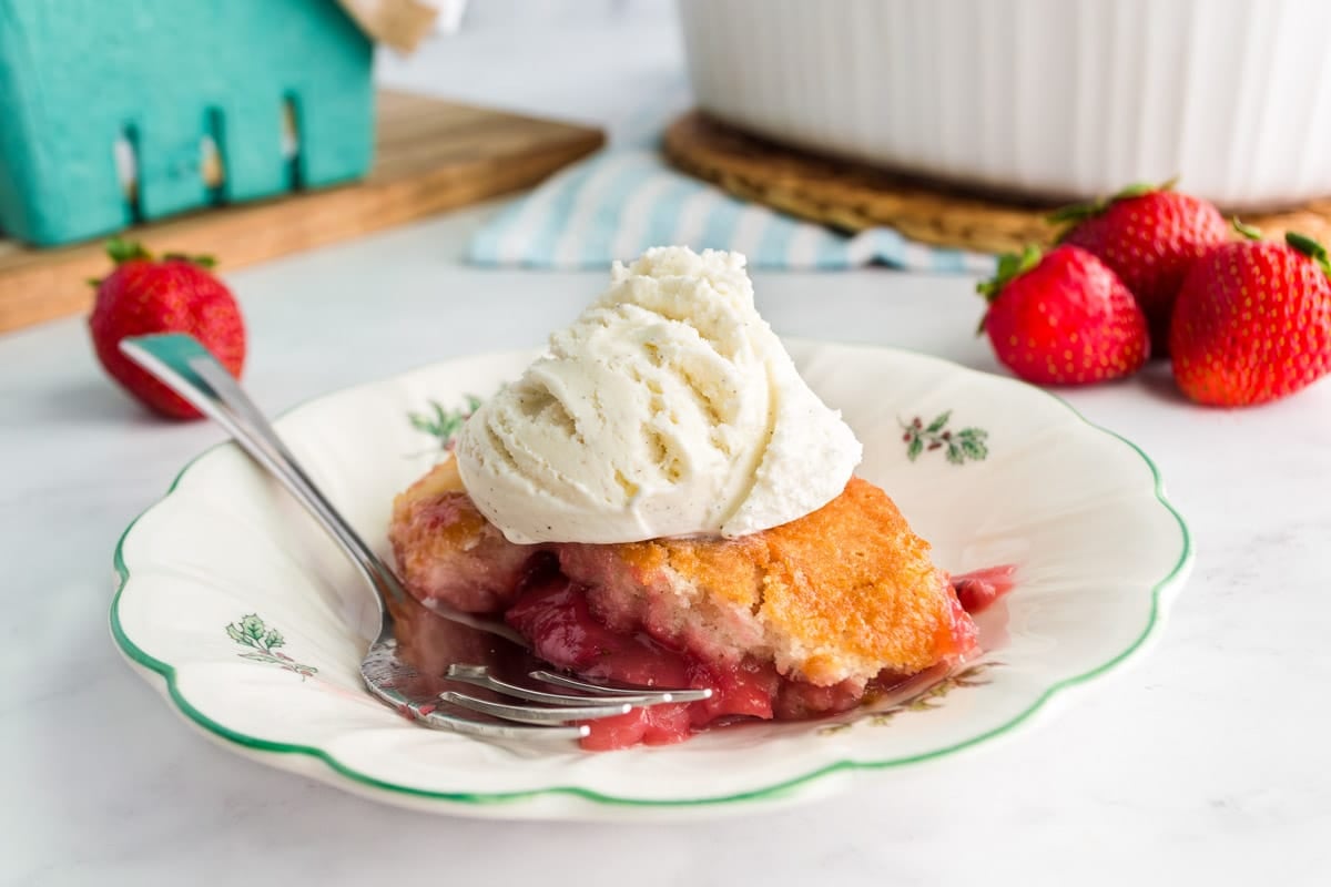 A plate with strawberry cobbler topped with a scoop of vanilla ice cream. A fork rests on the plate, and fresh strawberries are in the background.