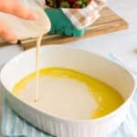 Pouring batter into a white baking dish with melted butter; strawberries in a container on a wooden board in the background.