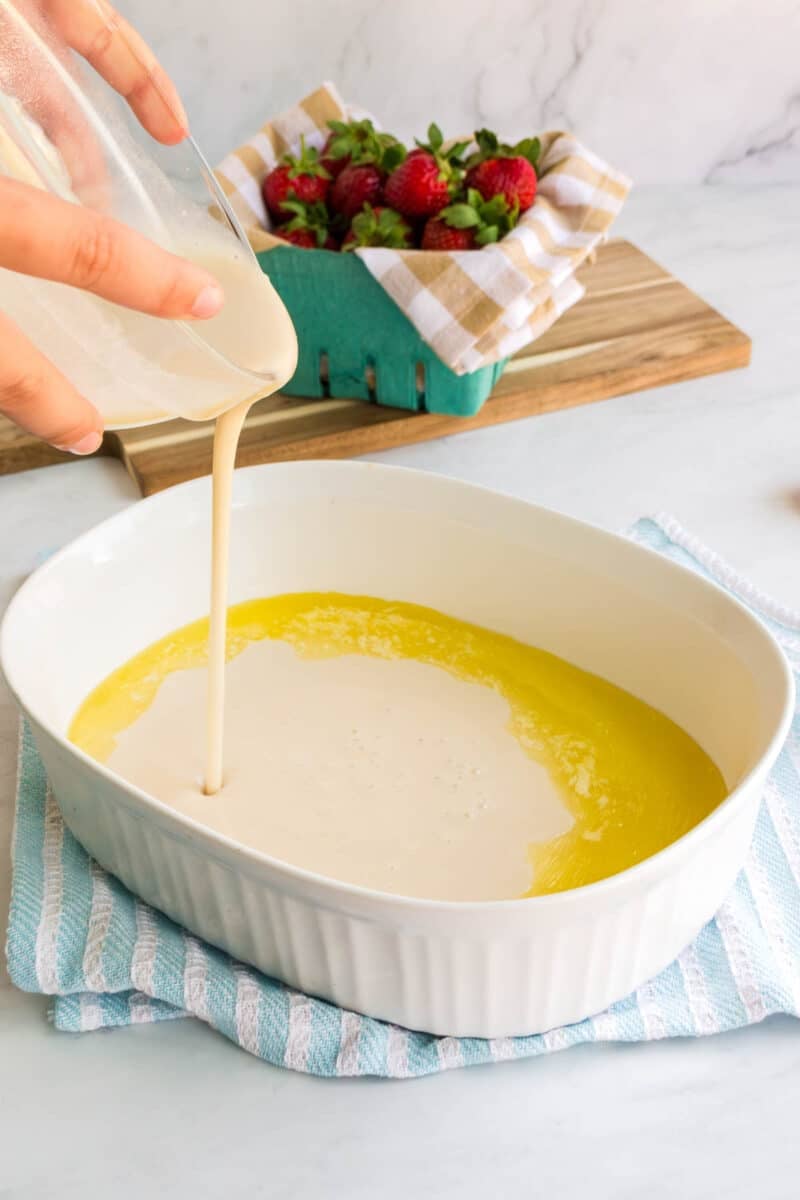 Pouring batter into a white baking dish with melted butter; strawberries in a container on a wooden board in the background.