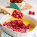 Wooden spoon scooping baked strawberries in a white dish, with fresh strawberries in a basket and wooden board in the background.