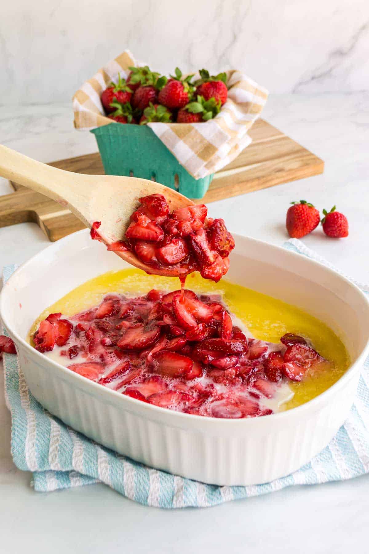 Wooden spoon scooping baked strawberries in a white dish, with fresh strawberries in a basket and wooden board in the background.