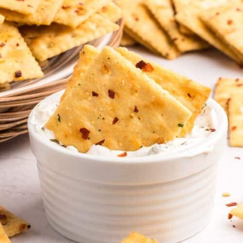Square cajun crackers topped with seasoning are dipped in a white creamy spread, with more crackers scattered and stacked in the background.