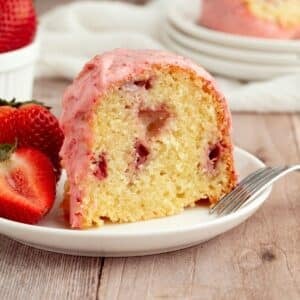 A slice of strawberry bundt cake with pink icing on a white plate, garnished with fresh strawberries, and a fork beside it.