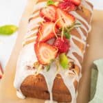 A strawberry loaf cake with white icing drizzled on top, garnished with sliced strawberries, lemon zest, and mint leaves, placed on parchment paper.