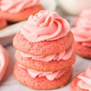 Three pink cookies stacked on top of each other, each with a swirl of pink frosting, placed on a white surface.