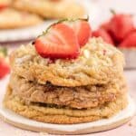 A stack of cookies with white chocolate chips, topped with halved fresh strawberries, sits on a round ceramic plate. Whole strawberries are in the background.