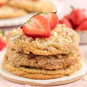 A stack of cookies with white chocolate chips, topped with halved fresh strawberries, sits on a round ceramic plate. Whole strawberries are in the background.