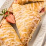 Three glazed strawberry turnovers are arranged on a rectangular white plate, with a sliced strawberry garnish on the side.