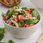 A white bowl filled with spinach salad featuring sliced strawberries, shredded chicken, walnuts, and crumbled cheese.