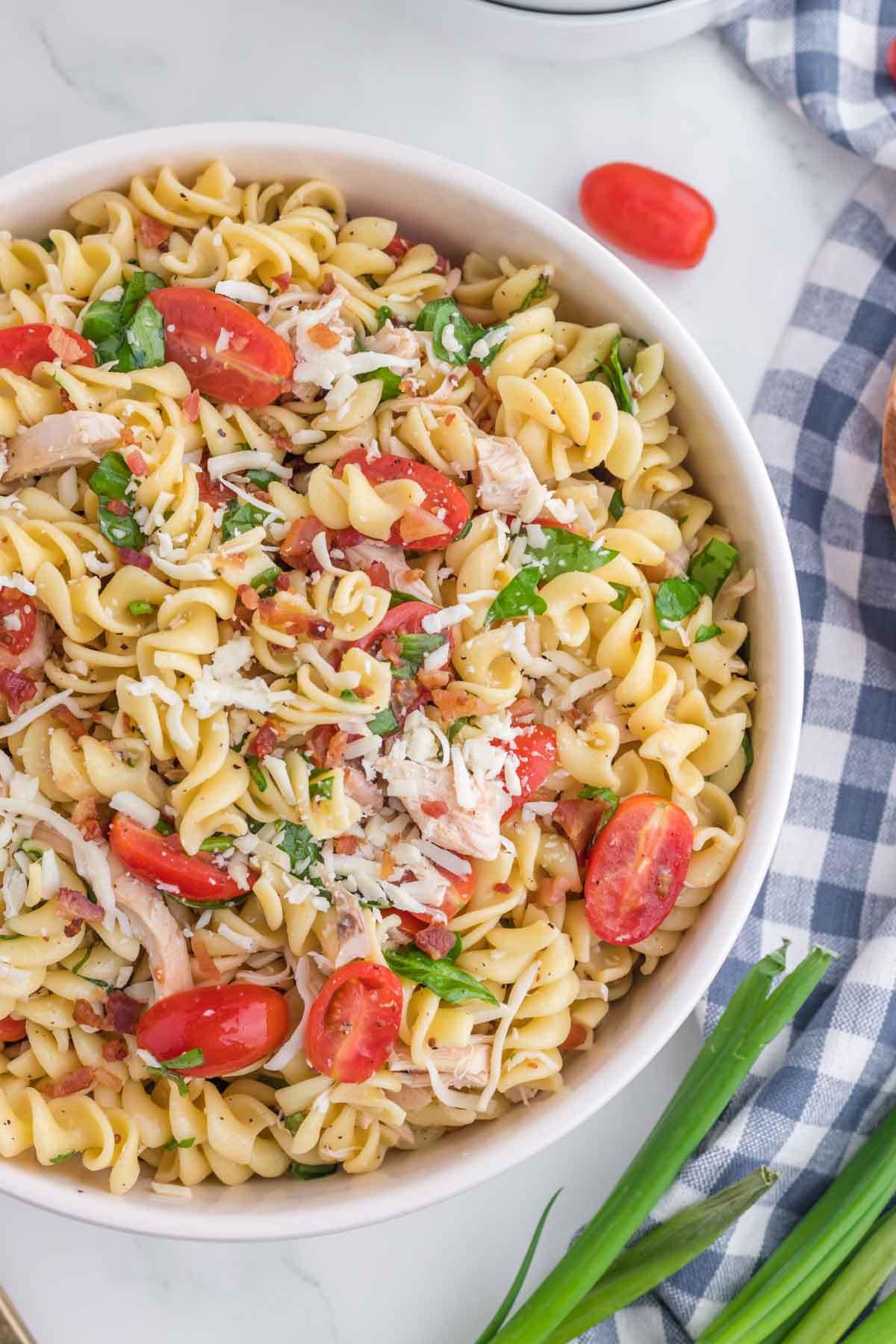 A bowl of rotini pasta salad with shredded cheese, cherry tomatoes, and chopped herbs on a marble surface next to a checkered cloth and green onions.