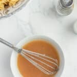 A bowl of brown sauce with a whisk inside, next to a bowl of cooked pasta, salt and pepper shakers on a white marble countertop.
