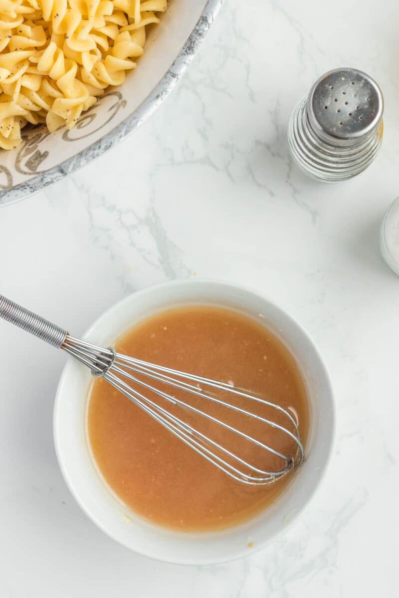 A bowl of brown sauce with a whisk inside, next to a bowl of cooked pasta, salt and pepper shakers on a white marble countertop.