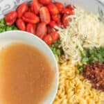 A bowl of dressing held above a salad containing cherry tomatoes, shredded cheese, rotini pasta, chopped greens, and bacon bits.