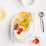 A plastic bag with uncooked pasta, flour, spices, and olive oil on a white surface, surrounded by small bowls and a metal measuring spoon.