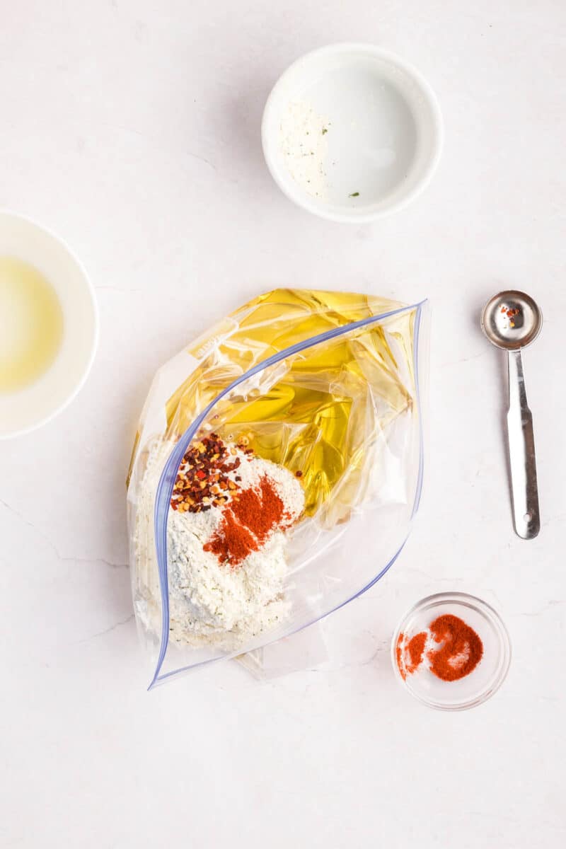 A plastic bag with uncooked pasta, flour, spices, and olive oil on a white surface, surrounded by small bowls and a metal measuring spoon.