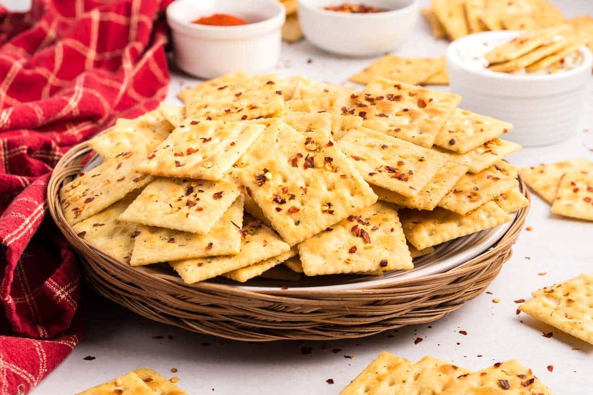 A wicker plate filled with square crackers topped with red pepper flakes, surrounded by more crackers and small bowls on a white surface.