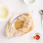 A plastic zip-top bag filled with marinade sits on a plate, surrounded by small bowls of ingredients and a metal measuring spoon on a white surface.