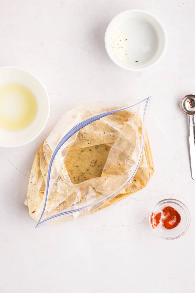 A plastic zip-top bag filled with marinade sits on a plate, surrounded by small bowls of ingredients and a metal measuring spoon on a white surface.