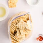 A ziplock bag filled with square crackers sits on a white surface, surrounded by small bowls containing oil, seasoning, and paprika.