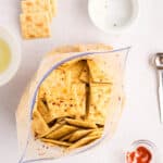 Opened plastic bag filled with seasoned saltine crackers on a white surface, surrounded by stacked crackers, spices in small bowls, and a measuring spoon.