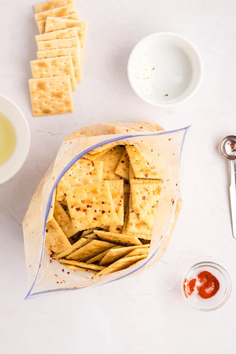 Opened plastic bag filled with seasoned saltine crackers on a white surface, surrounded by stacked crackers, spices in small bowls, and a measuring spoon.