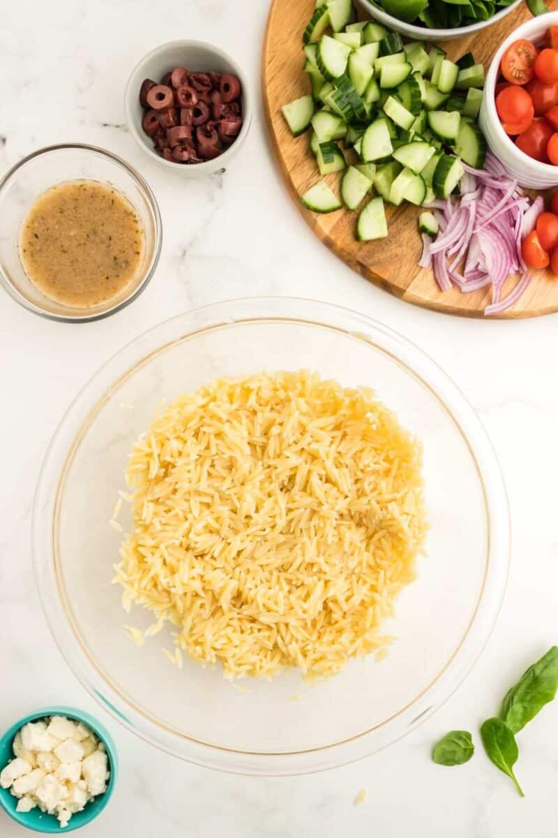 A glass bowl of cooked orzo surrounded by small bowls of vinaigrette, feta cheese, olives, and a platter of sliced cucumbers, tomatoes, spinach, and red onions.