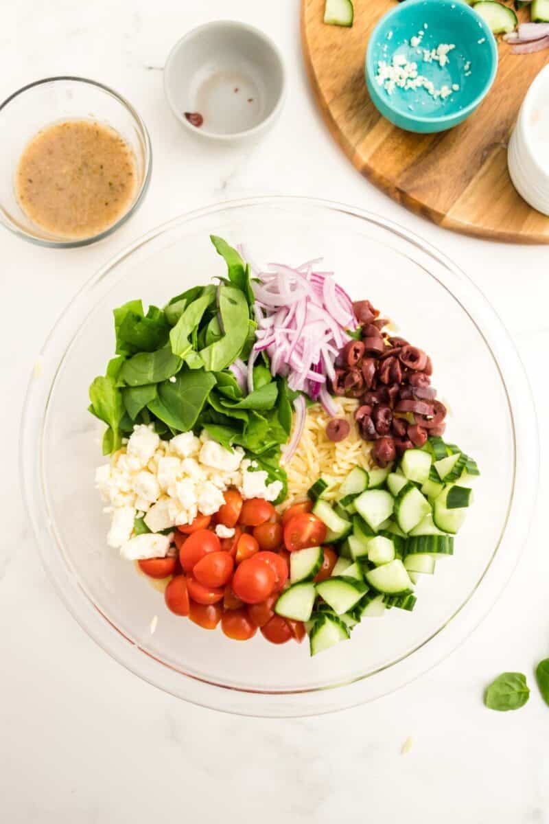 A glass bowl contains spinach, red onion, Kalamata olives, feta cheese, cherry tomatoes, and cucumber on a white surface with dressing and empty bowls nearby.