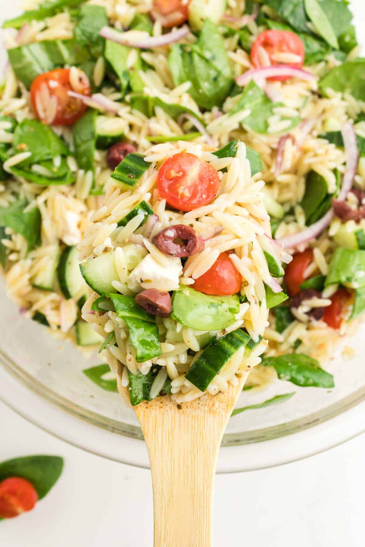 A wooden spoon holds a serving of orzo salad with tomatoes, cucumbers, spinach, red onions, kalamata olives, and feta cheese above a bowl of the same salad.