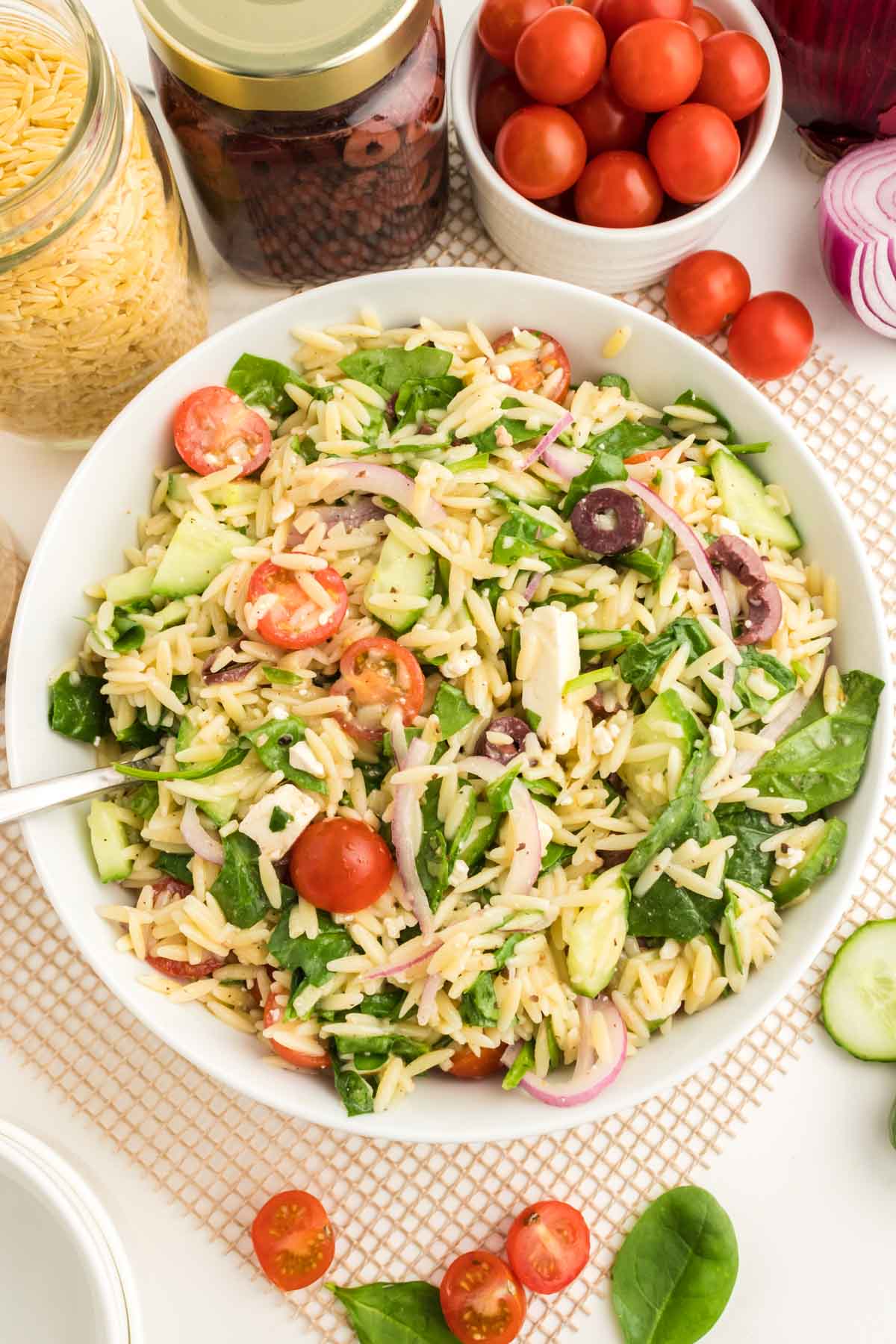 A bowl of Greek orzo salad with cherry tomatoes, cucumbers, red onions, spinach, olives, and feta cheese, surrounded by ingredients on a white surface.