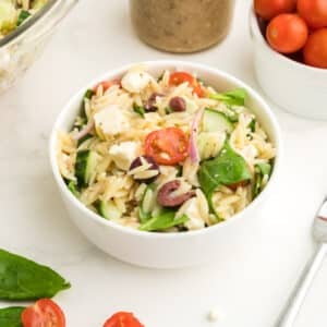 A bowl of Greek orzo pasta salad with spinach, cherry tomatoes, feta cheese, and olives sits on a white surface next to a fork, a bowl of cherry tomatoes, and fresh spinach leaves.