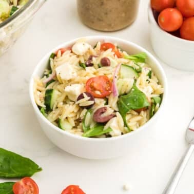 A bowl of Greek orzo pasta salad with spinach, cherry tomatoes, feta cheese, and olives sits on a white surface next to a fork, a bowl of cherry tomatoes, and fresh spinach leaves.