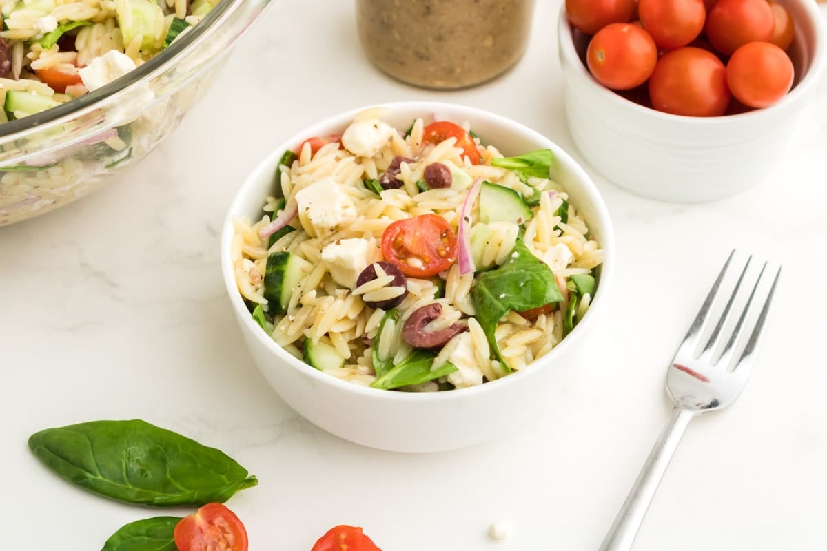 A bowl of Greek orzo pasta salad with spinach, cherry tomatoes, feta cheese, and olives sits on a white surface next to a fork, a bowl of cherry tomatoes, and fresh spinach leaves.