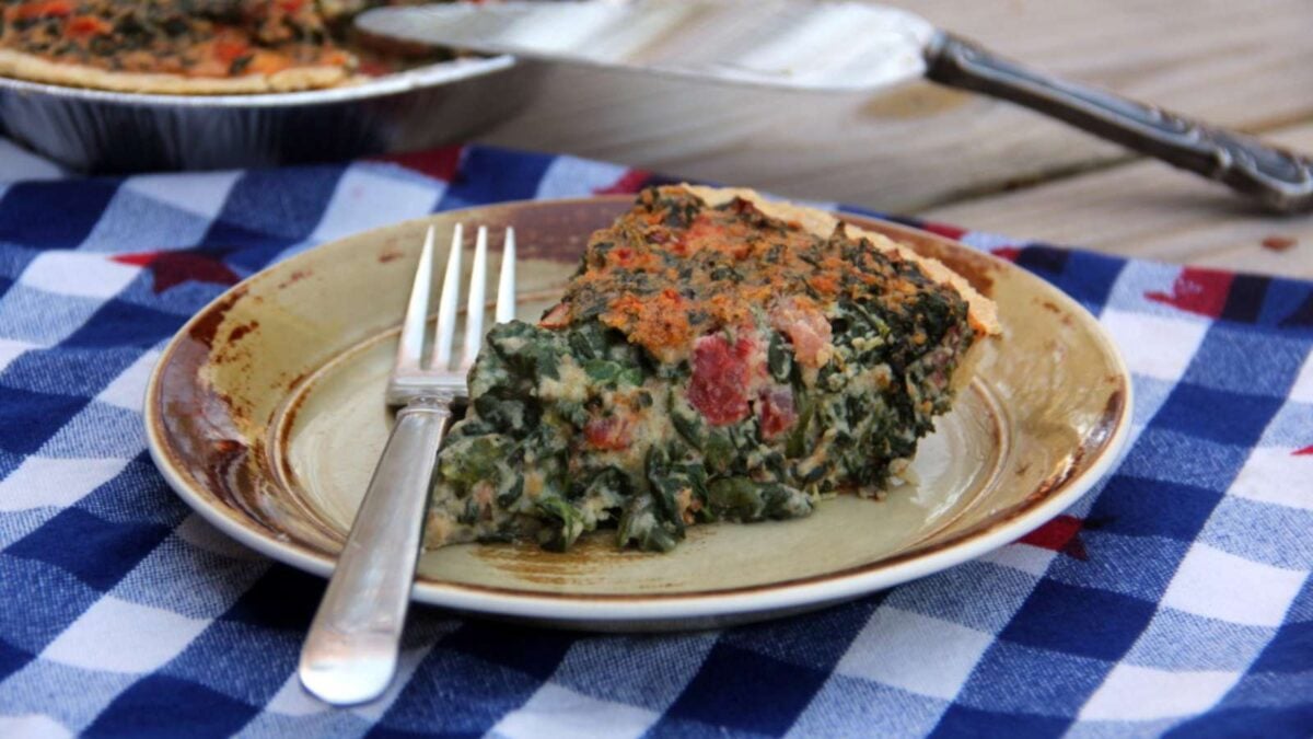 A slice of spinach and tomato quiche—one of our favorite healthy spinach recipes—is served on a plate with a fork, resting on a blue and white checkered cloth.