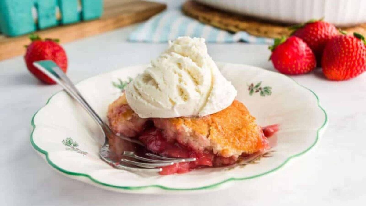 A serving of strawberry cobbler topped with a scoop of vanilla ice cream on a decorative plate, with a fork and fresh strawberries in the background—a classic treat from Southern cuisine.