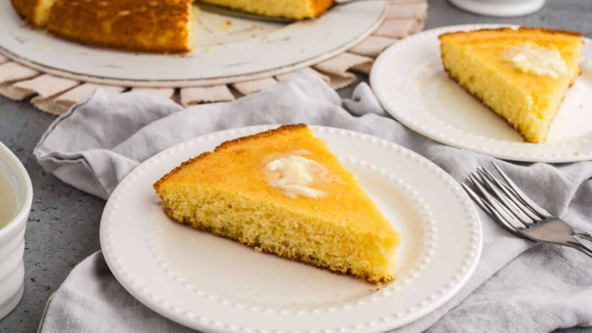 A slice of cornbread with a pat of melting butter sits on a white plate, showcasing the comfort of Southern cuisine recipes, with another slice and a serving fork in the background.