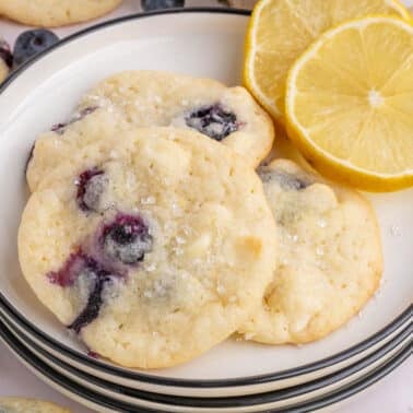 A plate of blueberry and white chocolate cookies is shown with two lemon slices, surrounded by more cookies and white chocolate chips.